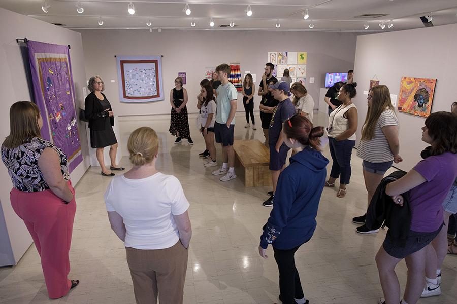 A group of students are gathered around a faculty member who is standing next to a large piece of artwork hanging on a gallery wall.