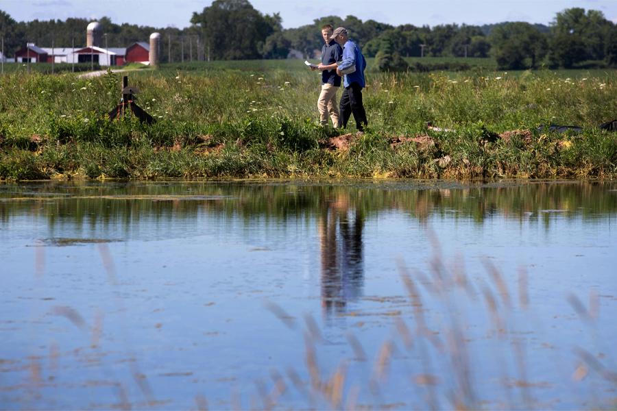 Two people walk along a waterway with a farm in the background.