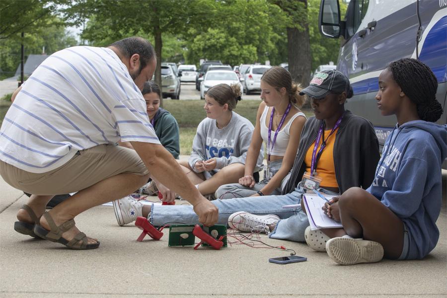 A faculty member sits with students outdoors.