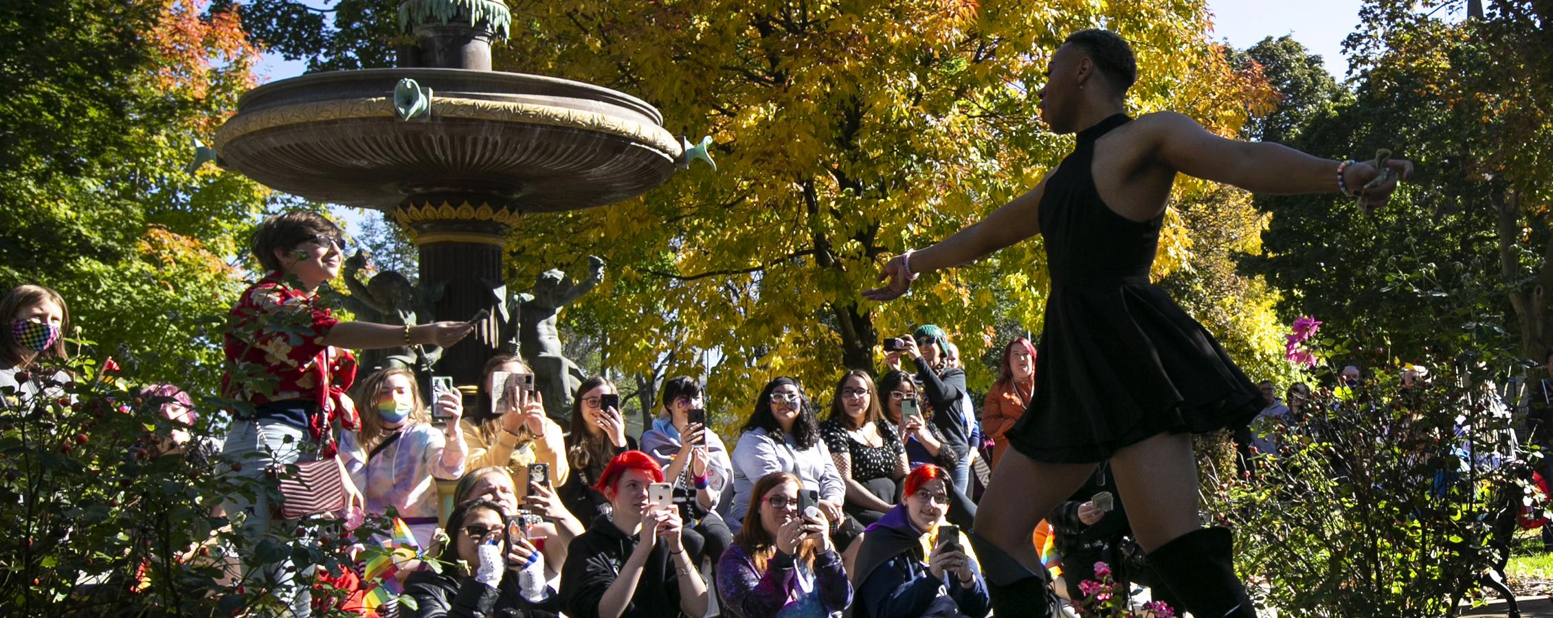 A person dances outdoors in front of a crowd by a water fountain.