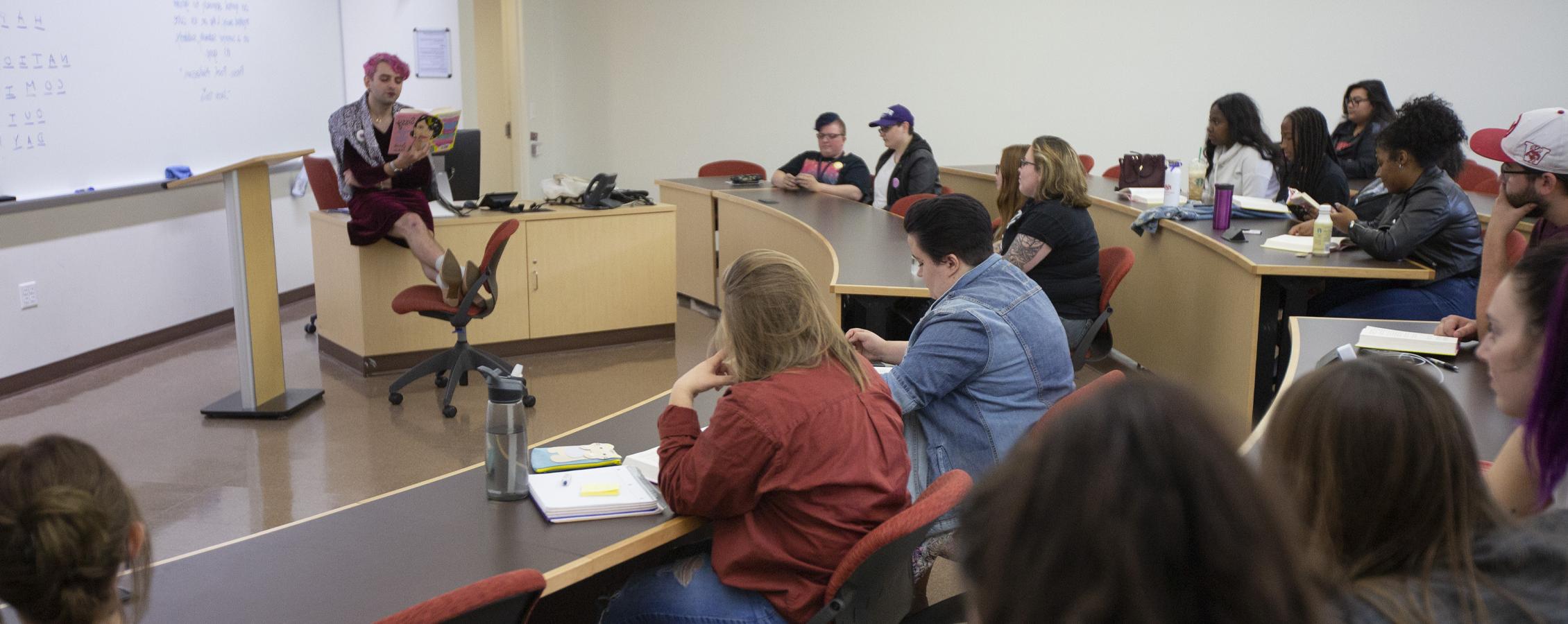 A person with pink hair reads from a book at the front of class.