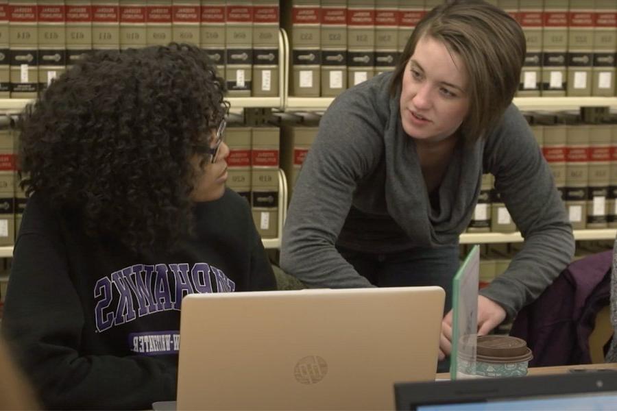 Two students discuss over books and a laptop.